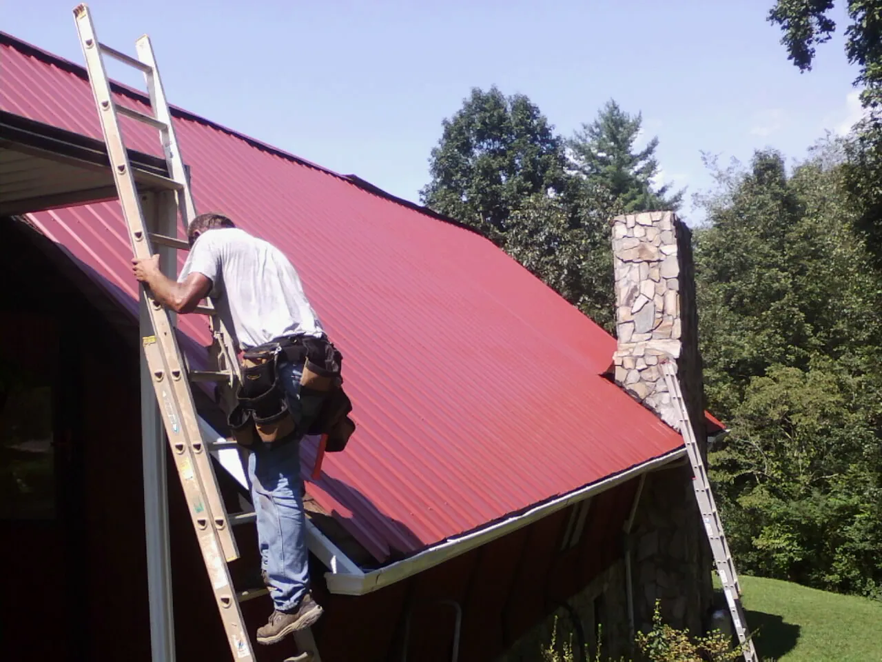 Roofer on a ladder beside a red metal roof and stone chimney — Soffit & Fascia Repair by Roofing Brothers in Portal